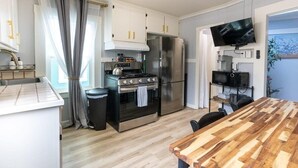 A compact kitchen with a stove, oven, and refrigerator, a wooden dining table, and a TV mounted on the wall. - Capitola Village Bungalow - 2 Blocks from the Sand (Capitola)
