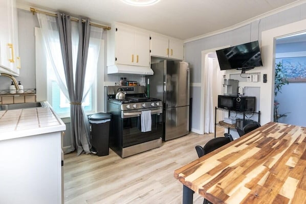 A compact kitchen with a stove, oven, and refrigerator, a wooden dining table, and a TV mounted on the wall.