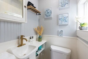 Een badkamer met een witte wastafel, een gouden kraan en een toilet. Er hangen kunstwerken in lijsten aan de muur en een klein schapje met toiletartikelen.