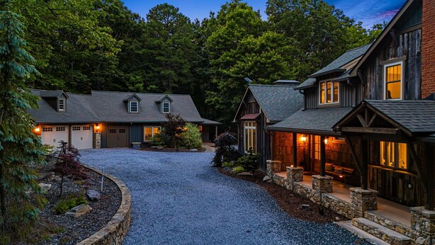 A two-story house with a gravel driveway, surrounded by trees and featuring a stone border.