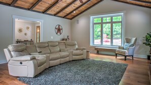 A spacious living room with a large sectional sofa, a ceiling fan, a large window with a view of greenery, and a wooden ceiling.
