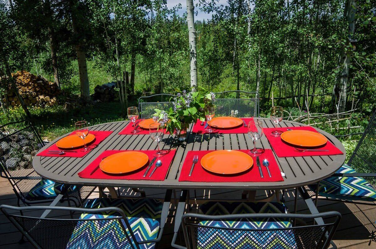 A round outdoor dining table set with orange plates, red placemats, and metal chairs with patterned cushions.