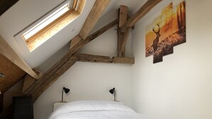Un dormitorio con techo inclinado, vigas de madera, una cama, dos lámparas de noche, una silla y un cuadro de un ciervo.