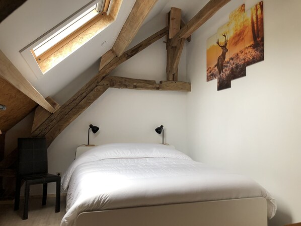 A bedroom with a sloped ceiling, wooden beams, a bed, two bedside lamps, a chair, and a painting of a deer. - Propriété de Campagne Rénovée Avec Barbecue, à Proximité Sentiers de Randonnées (Sivry-Rance)