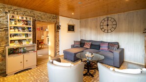 Una sala de estar acogedora con pared de piedra, techo de madera, un sofá gris con cojines estampados, una mesa de centro y una estantería llena de libros y revistas.