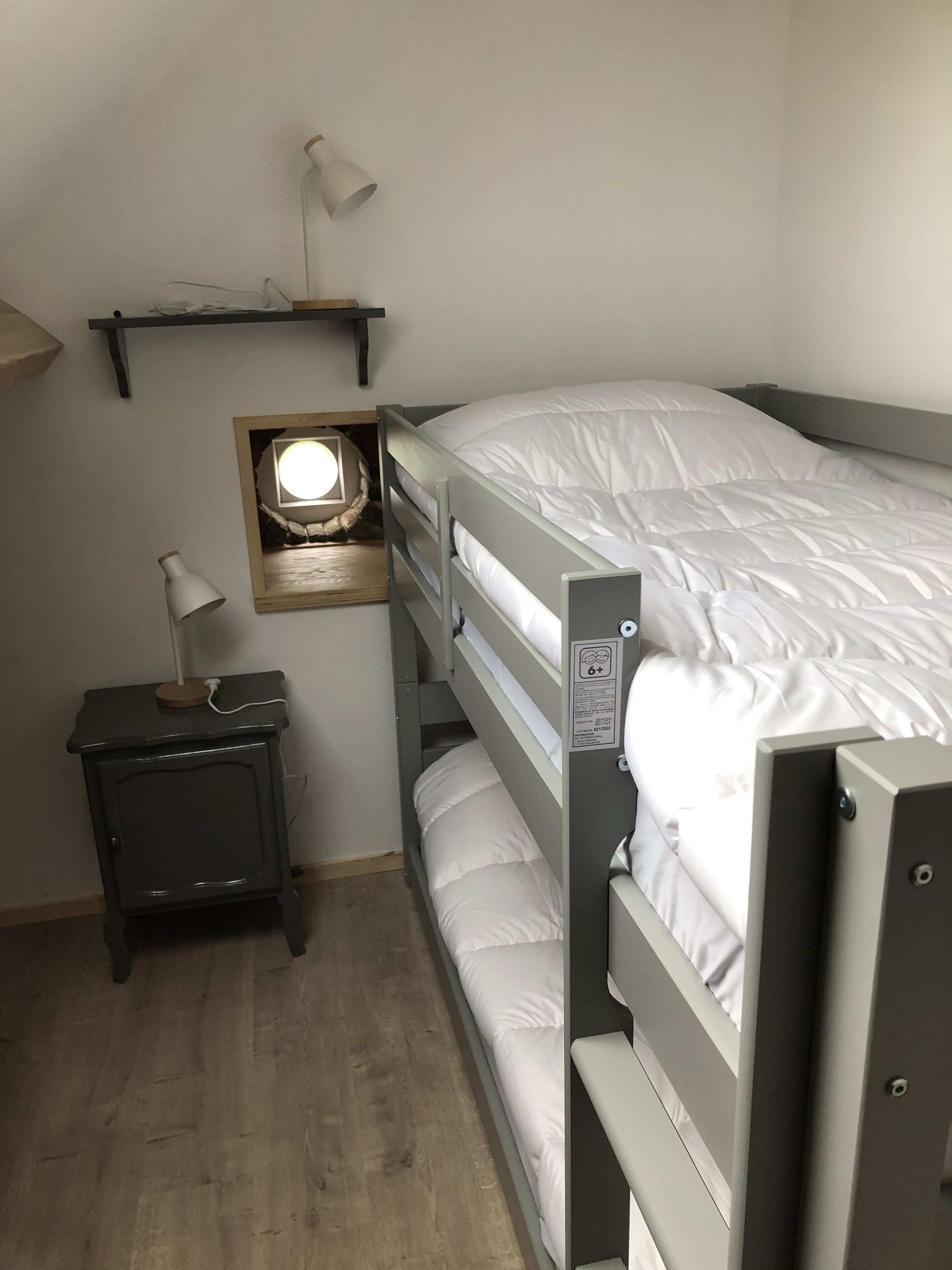 Una cama individual superpuesta con una mesita de noche y una lámpara, un estante encima de la cama y una pequeña ventana con una luminaria.