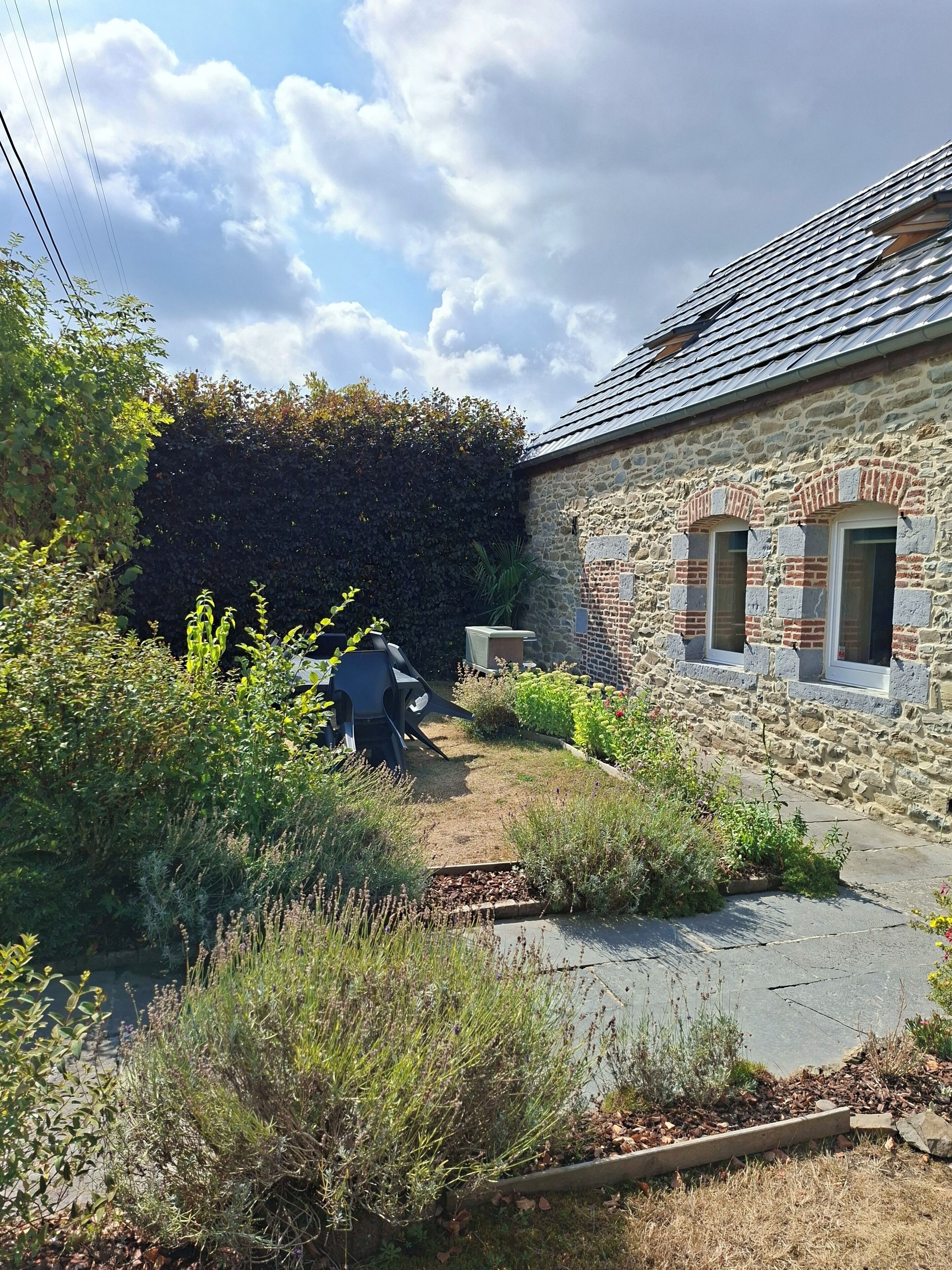 Een stenen gebouw met een schuin dak, een tuin met verschillende planten en een pad dat naar de ingang leidt.