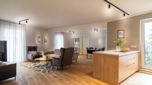 A spacious living room with a sofa, armchair, coffee table, and a wooden island with a sink. There is a dining area with a table and chairs, and a kitchen area with cabinets and a window. - Sleep Inn Medienhafen Suites (Düsseldorf)