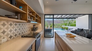 A modern kitchen with wooden shelves, a built-in stove, and a large window overlooking a pool and greenery. - Modern Villa Overlooking Lush National Park (Huatulco)