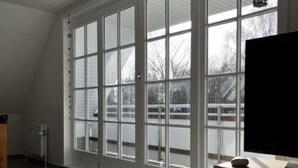 Una habitación amplia con ventanas grandes, un televisor de pantalla plana sobre un soporte de madera y un radiador en la pared.
