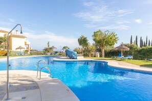 A swimming pool with lounge chairs and umbrellas, surrounded by greenery and a clear sky.