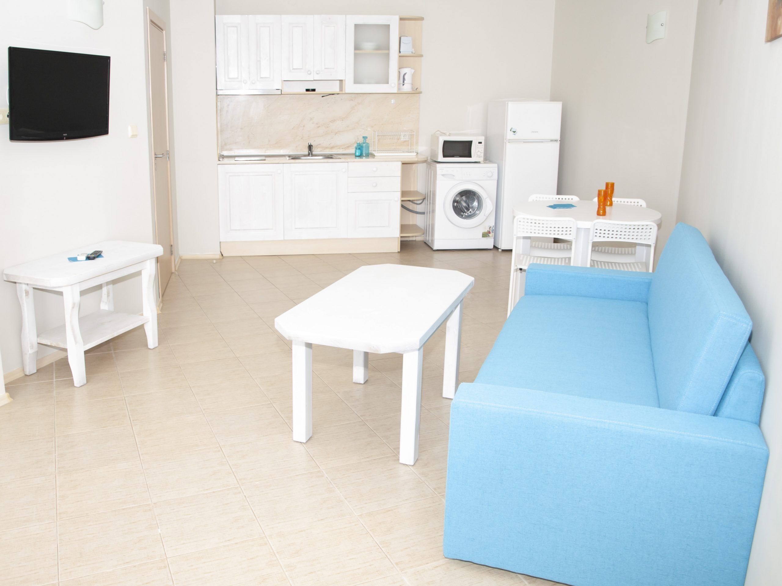 A compact living space with a blue sofa, a white table, a small white table, a washing machine, a microwave, and a TV mounted on the wall.