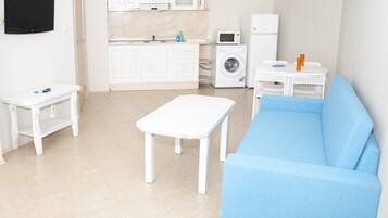 A compact living space with a blue sofa, a white table, a small white table, a washing machine, a microwave, and a TV mounted on the wall.