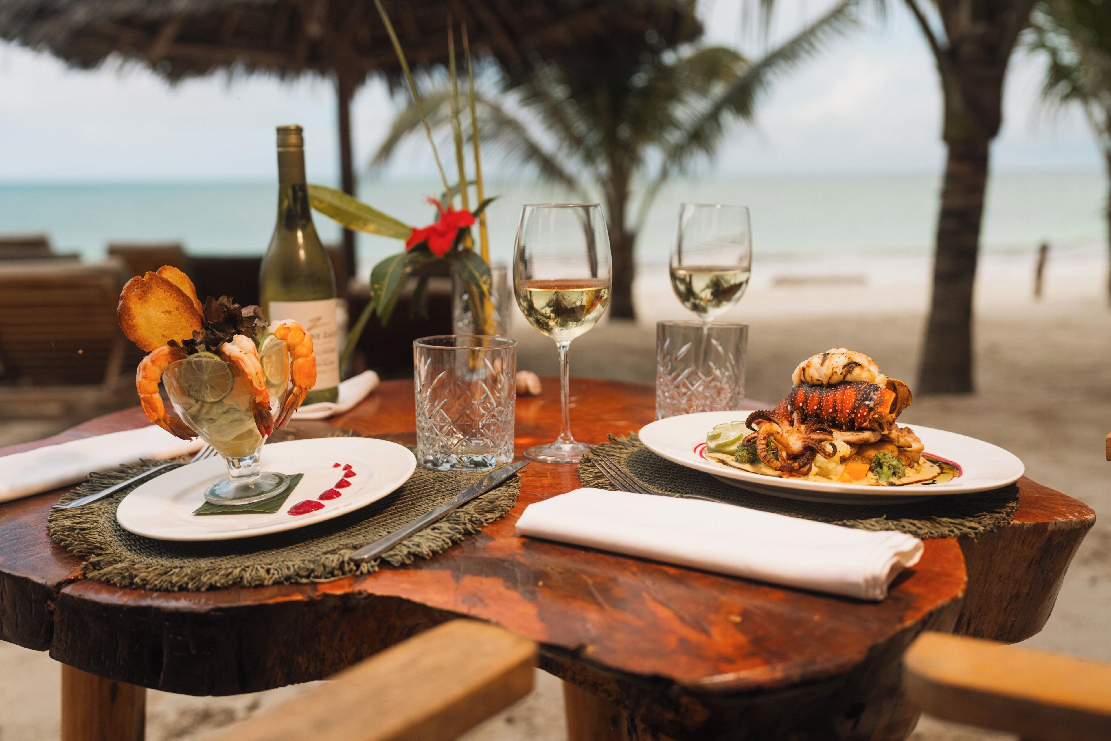 Une table en bois, dressée pour un repas, avec deux verres à vin et une bouteille de vin, le tout installé sur une plage.