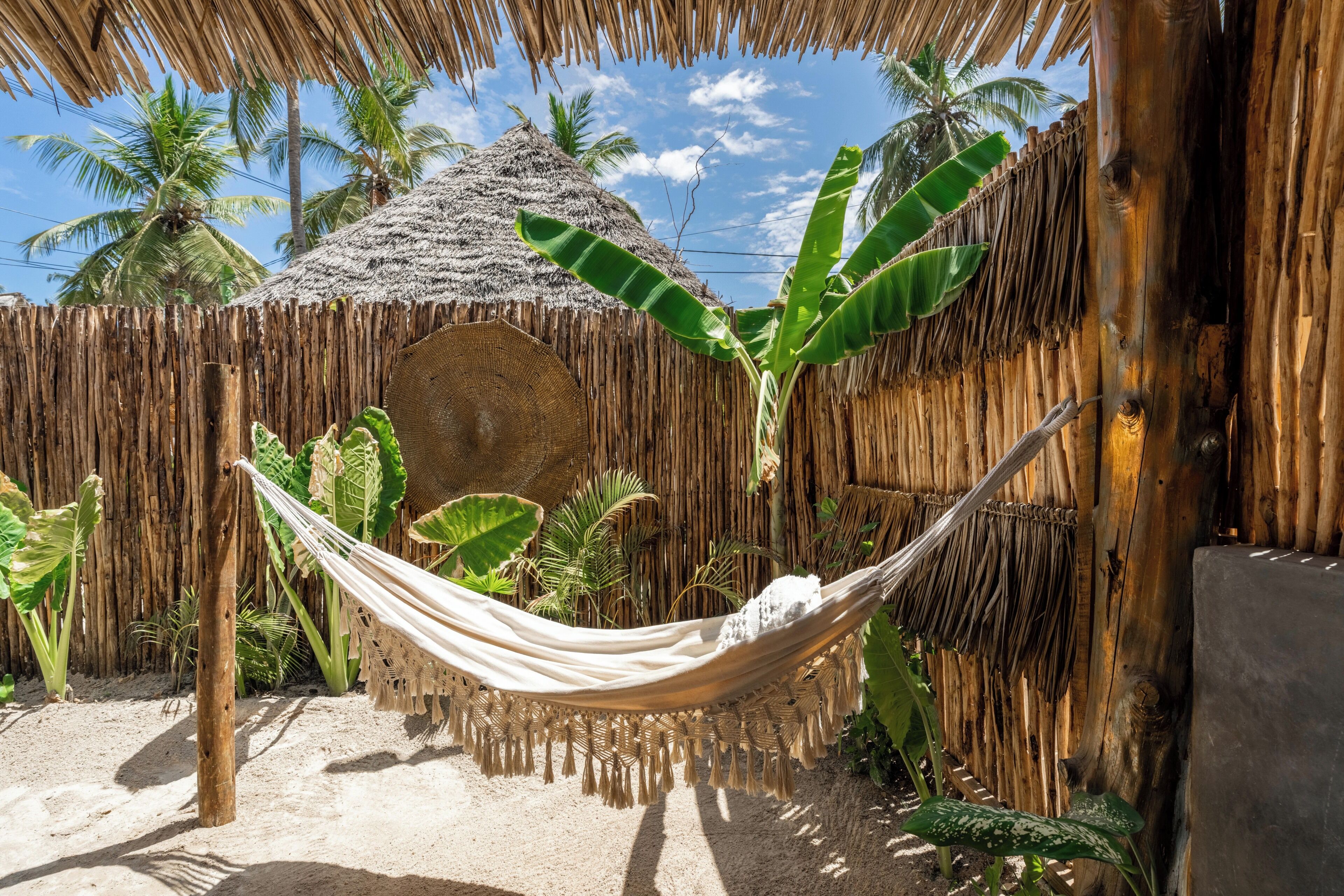 Un hamac dans un cadre tropical, avec un toit de chaume, des palmiers et un sol sablonneux.