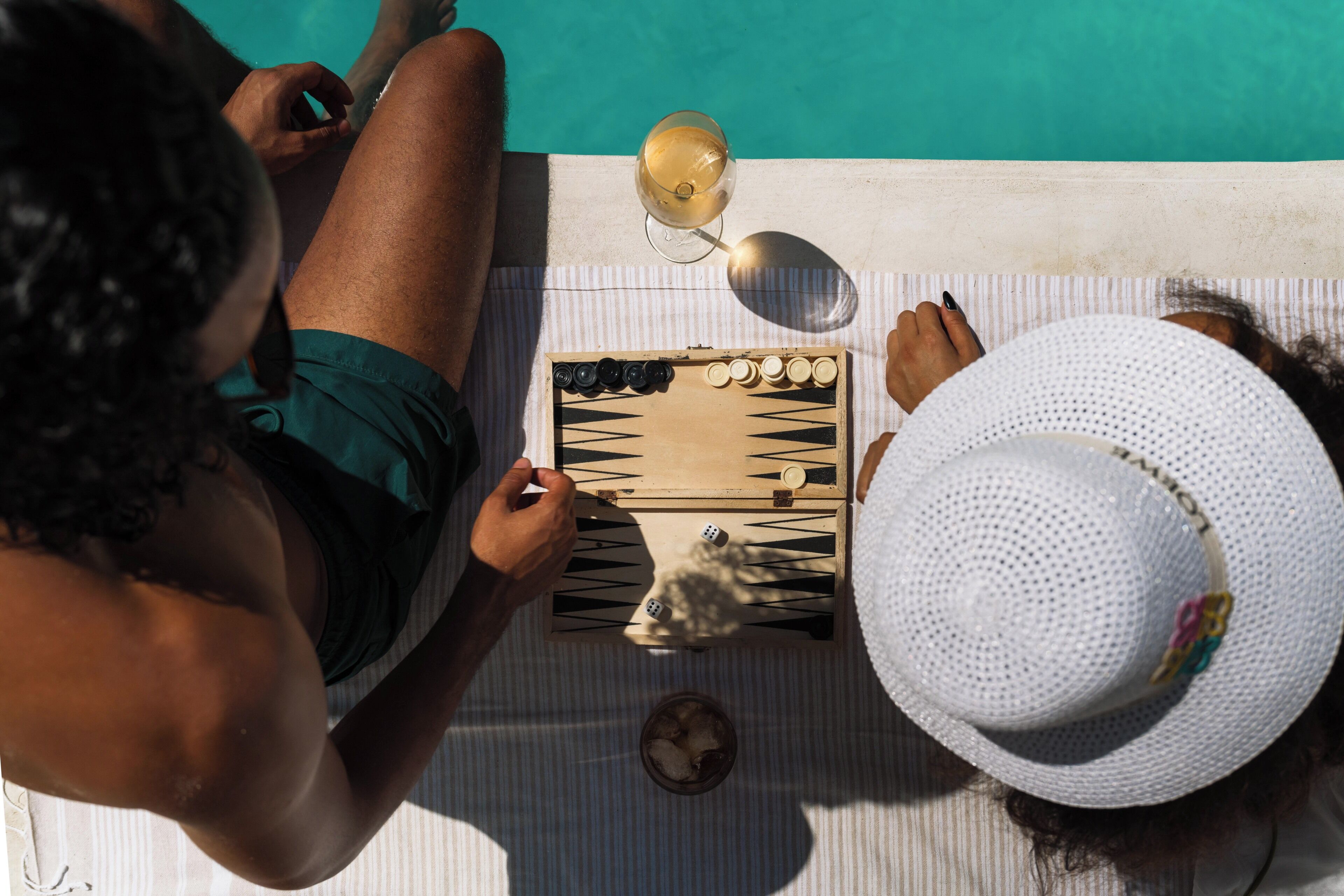 Two people playing a board game on a striped towel by a poolside.