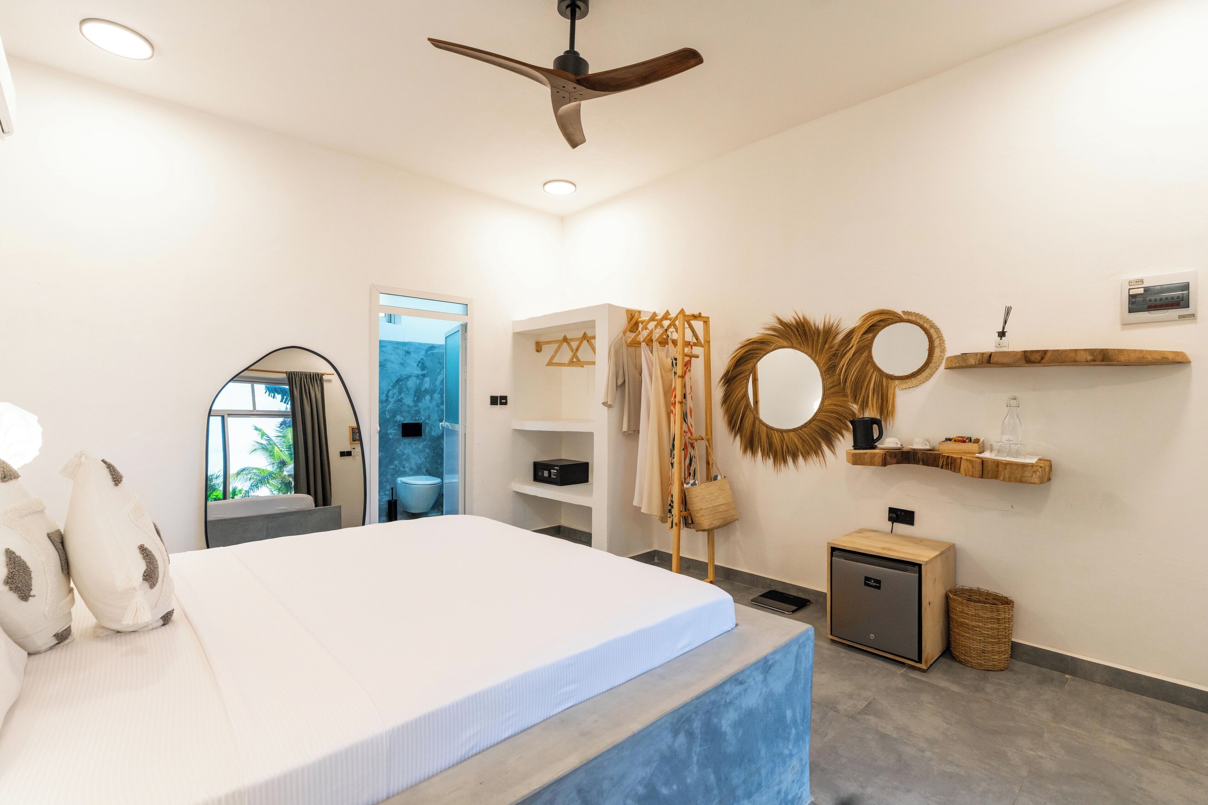 A modern hotel room with a large bed, a ceiling fan, a small cabinet, a mirror, and a view of the outdoors.