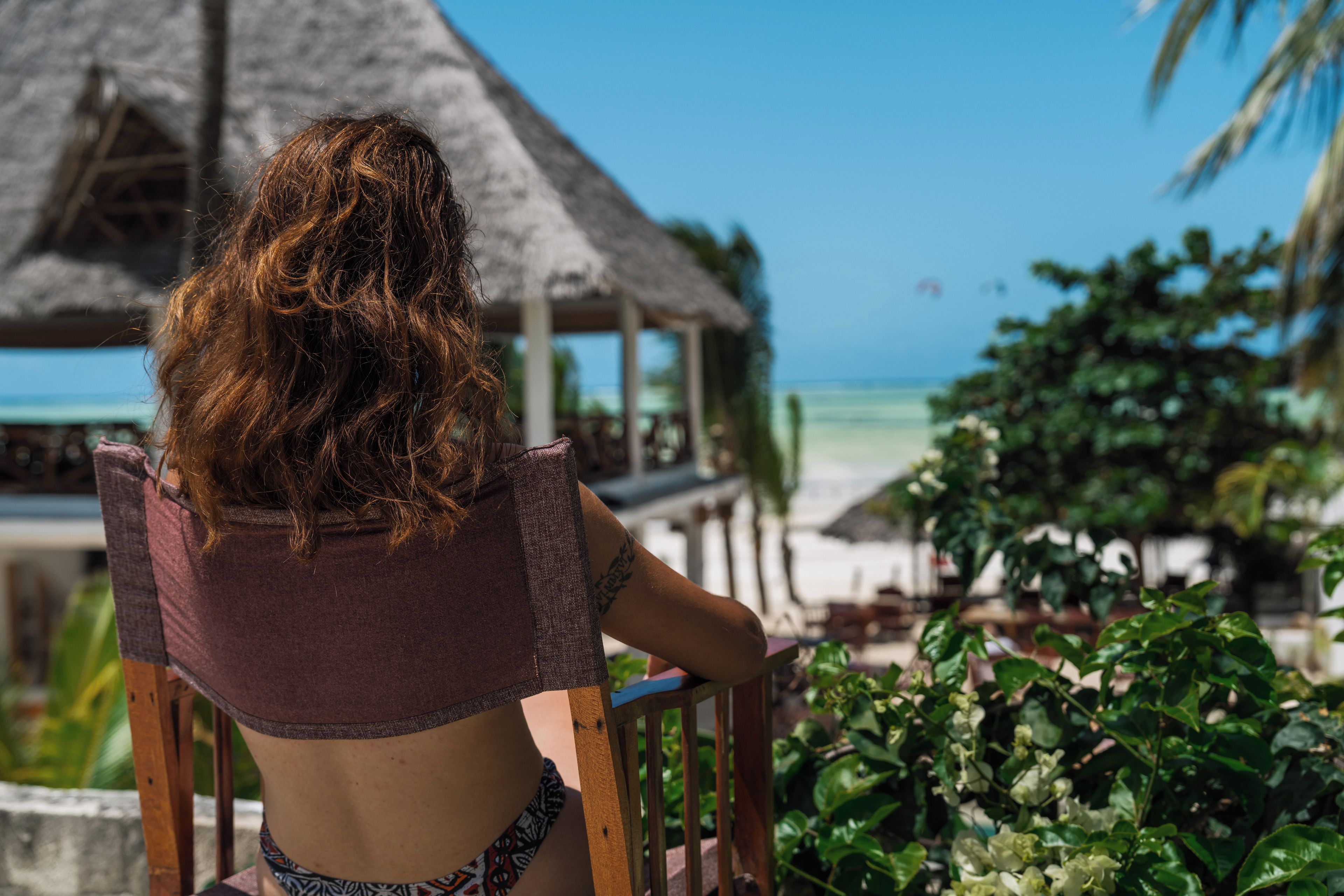 Eine Person sitzt an einem Holztisch mit Blick auf einen Strand und ein Gebäude mit einem Reetdach.
