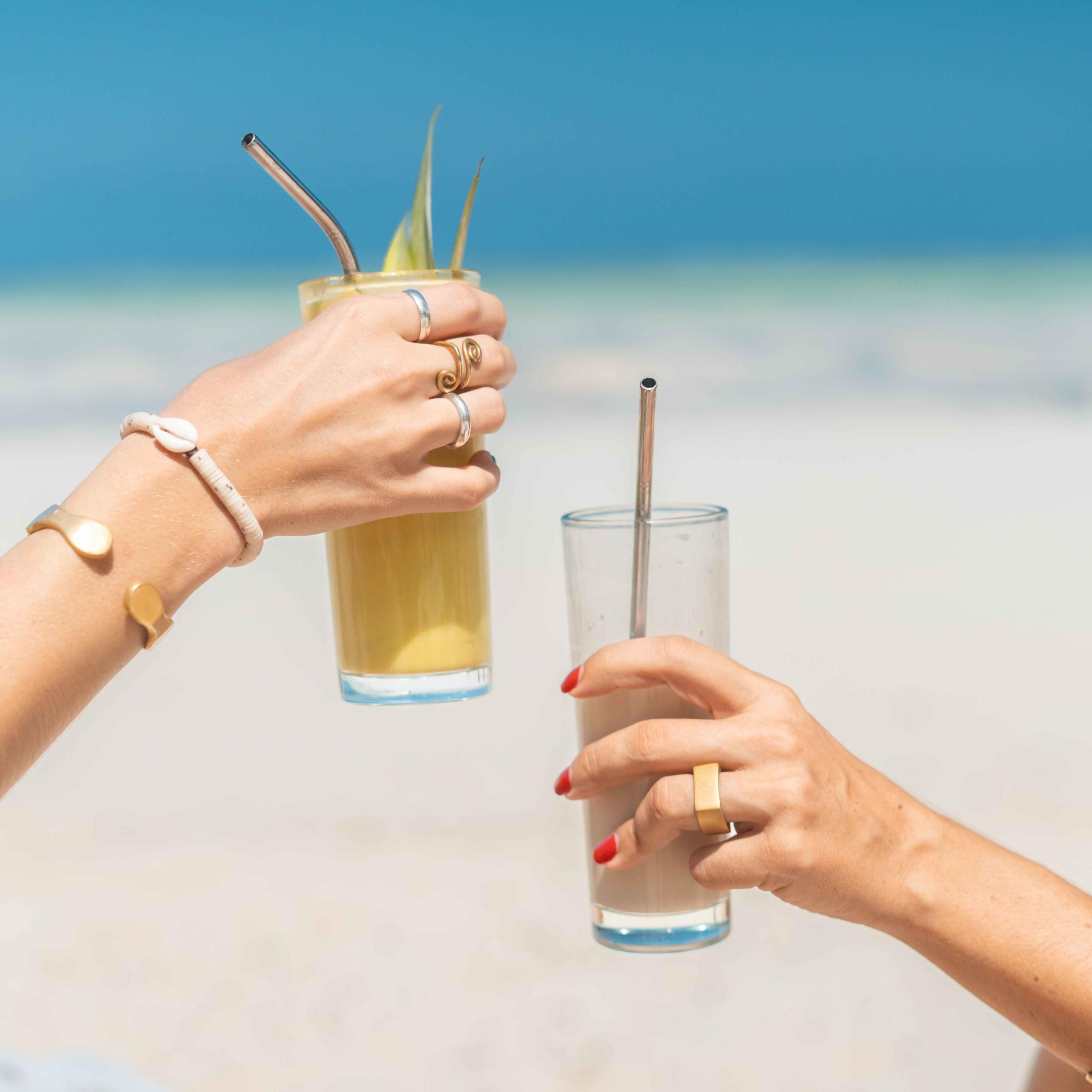 Dos manos sosteniendo vasos con pajitas en una playa.