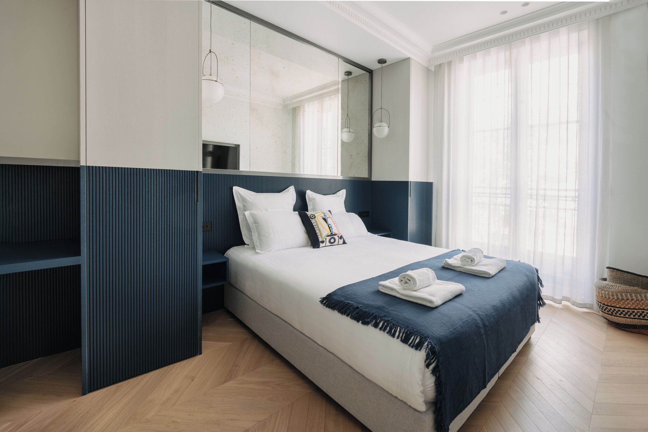 A modern hotel room with a large bed, a wardrobe, a mirror, and a window with sheer curtains.