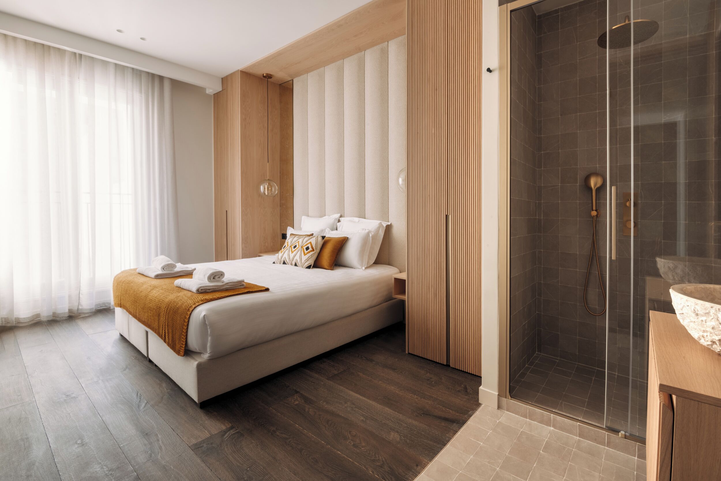 A modern hotel room with a large bed, a wooden wardrobe, a glass shower enclosure, and a wooden vanity.