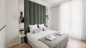 A modern hotel room with a large bed, a nightstand, a mirror, and a window with sheer curtains.