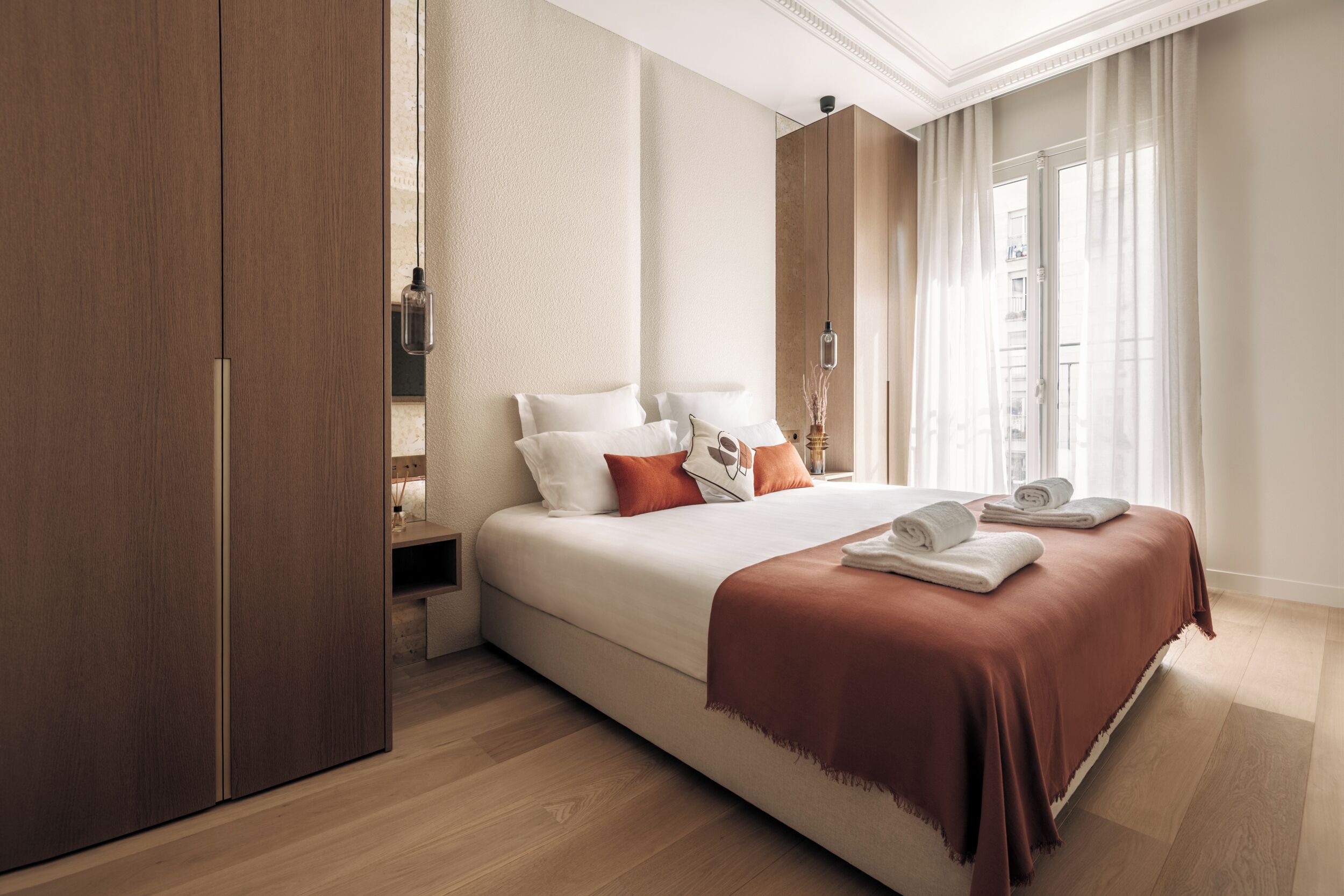 A modern hotel room with a large bed, a wardrobe, a nightstand, and a window with curtains.
