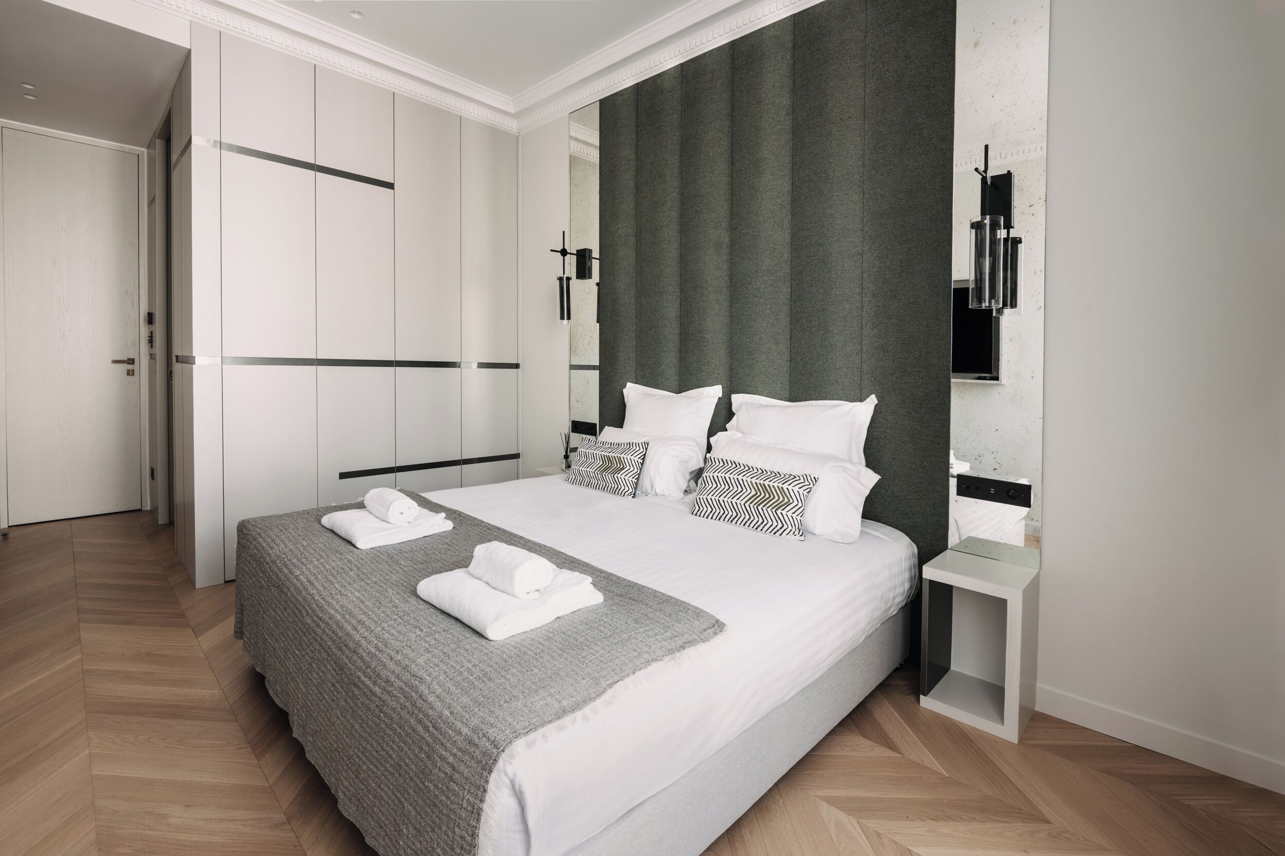Una habitación de hotel moderna con una cama grande, ropa de cama gris y un cabecero gris. Hay una mesita de noche pequeña con una lámpara, un armario y un espejo en la pared.