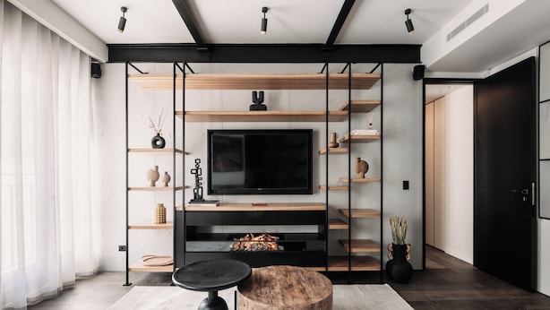 A modern living room with a large flat-screen TV, wooden shelves, a round coffee table, and a black-framed sofa.