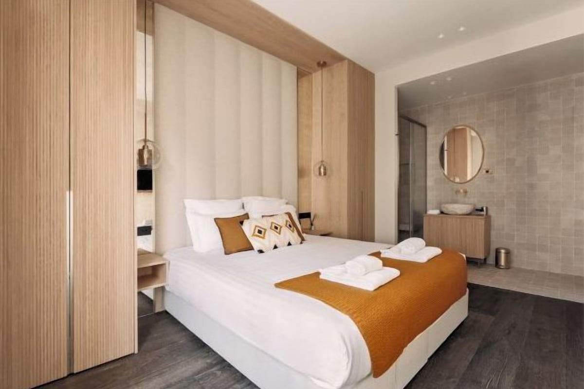A modern hotel room with a large bed, wooden wardrobe, and a bathroom with a round mirror and sink.