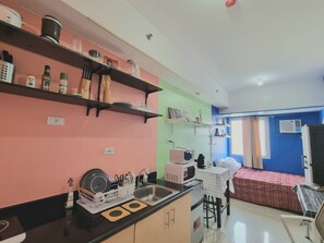 A compact kitchen with a sink, countertop, and shelves with kitchenware. A dining area with a table and chairs is visible in the background.