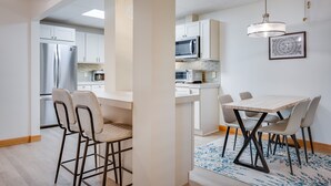 Une cuisine moderne avec des armoires blanches, un réfrigérateur en acier inoxydable et un four à micro-ondes. On y trouve un coin repas avec une table et des chaises, ainsi qu’un tapis à motifs sur le sol.
