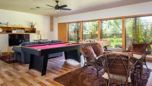 Un salon équipé d’une table de billard, d’un baby-foot, de fauteuils en osier, d’un ventilateur de plafond et de grandes fenêtres donnant sur l’extérieur.