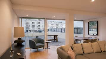 Ein modernes Wohnzimmer mit einer Couch, einem Tisch und einem Sessel. Ein Balkon mit Geländer bietet einen Blick auf Gebäude.