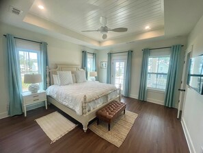 A bedroom with a bed, a nightstand, a bench, a ceiling fan, and large windows with curtains.