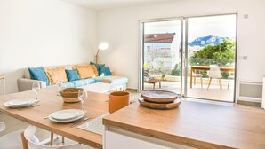 A modern living room with a wooden dining table set for four, a sofa with colorful cushions, and a balcony with a view of mountains and buildings. - Bail mobilité - Appart 2 ch parking et terrasse (Marseille)