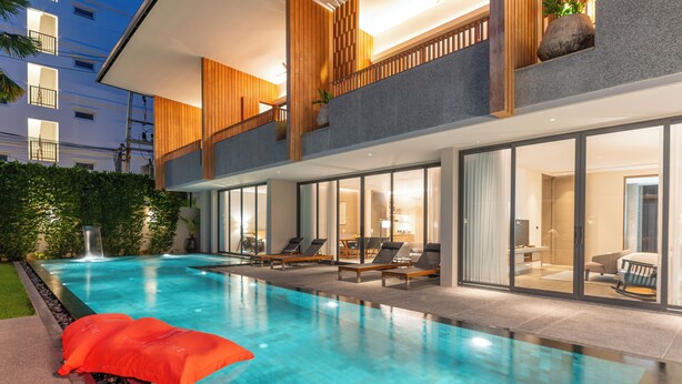 A modern hotel with a large outdoor swimming pool, a second-floor balcony with wooden paneling, and a well-lit interior. - Villa De Time The Luxury Private Pool Villa (Pattaya)