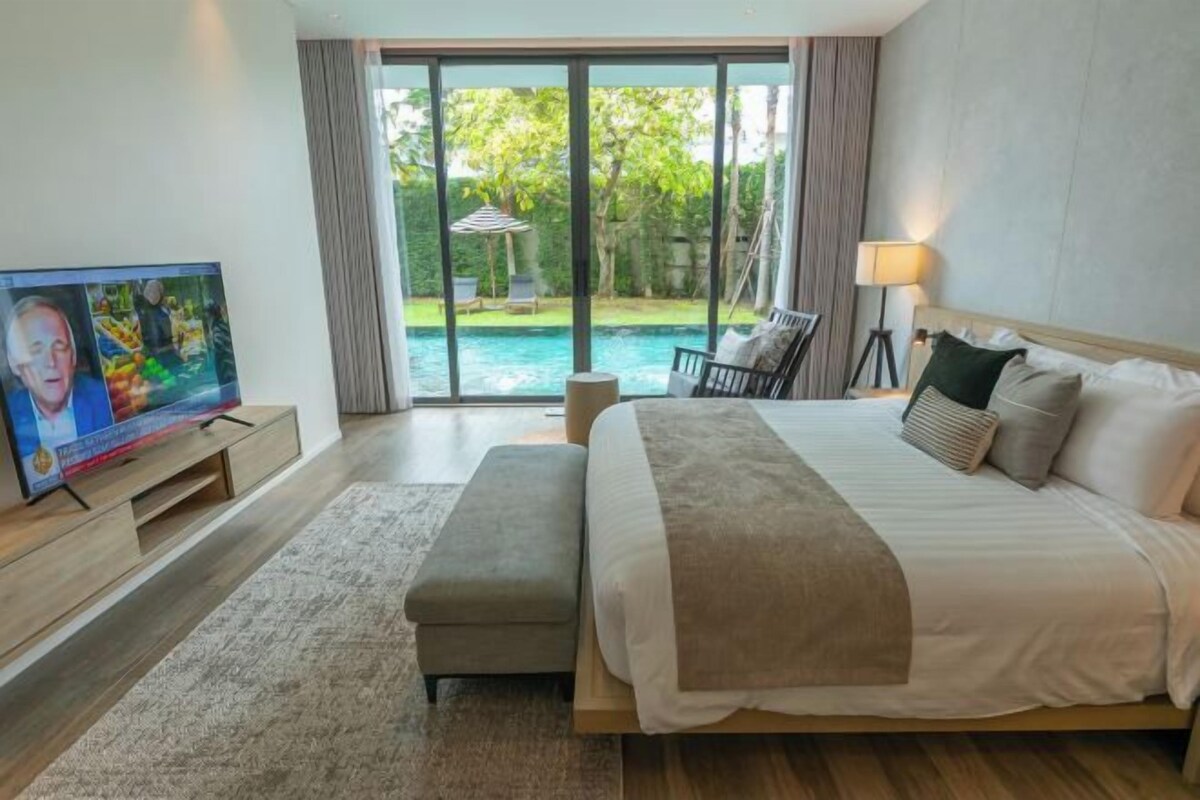 A hotel room with a large bed, a TV on a wooden stand, a bench, and a view of a swimming pool and garden.