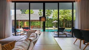 A modern living room with a large sofa, a wooden coffee table, and a view of a swimming pool and outdoor seating area through large glass doors. - Villa De Time The Luxury Private Pool Villa (Pattaya)