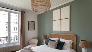 A neatly arranged bedroom with a large bed, two bedside tables with lamps, a chair, a window with curtains, and artwork on the wall.