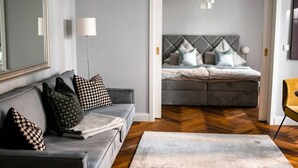 Ein modernes Wohnzimmer mit grauem Sofa, gemusterten Kissen, Holzboden und einem Bett mit grauem Kopfteil im Hintergrund.