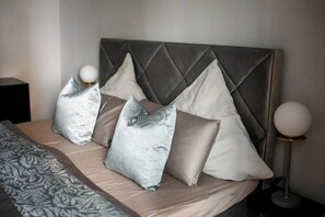 A bed with a grey upholstered headboard, multiple pillows, a patterned throw, and a bedside lamp.