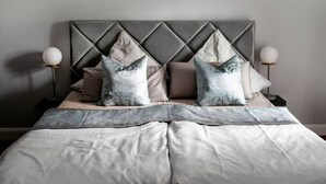 Ein Bett mit grauem Kopfteil, zwei weiße Nachttischlampen und eine Bettdecke in Grau und Weiß mit Muster.