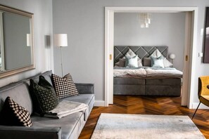 A modern living room with a grey sofa, patterned cushions, a large mirror, and a view into a bedroom with a grey upholstered bed and a herringbone-patterned floor.
