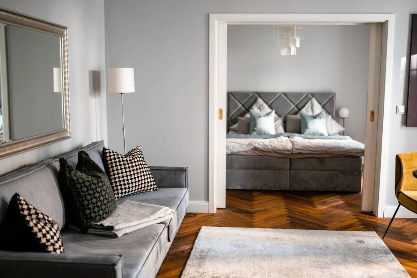 A modern living room with a grey sofa, patterned cushions, a large mirror, and a view into a bedroom with a grey upholstered bed and a herringbone-patterned floor.