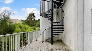 A spiral staircase on a wooden deck with a white railing, leading to a building with a white exterior.