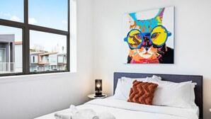 A hotel room with a bed, a window, a painting of a cat, and a nightstand with a lamp.