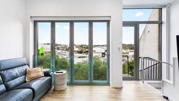A modern living room with a large sliding glass door leading to a balcony, a black leather sofa, and a small round table.