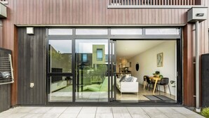 A modern house with a sliding glass door entrance leading to a living area with a sofa, table, and chairs. - Hobsonville Point Enchanter with Parking and Wi-fi (Auckland West)
