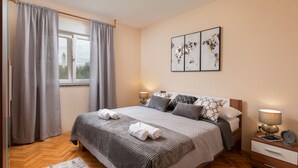 Une chambre à coucher bien rangée, avec un lit, des lampes de chevet, une œuvre d’art accrochée au mur et une fenêtre avec des rideaux.